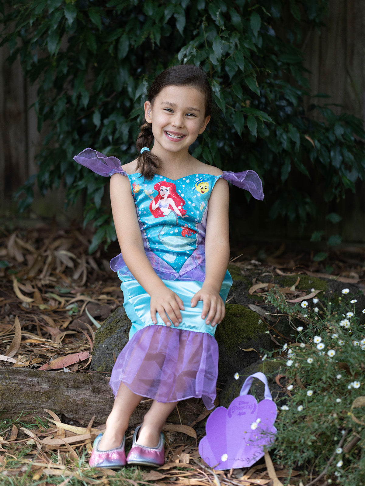 Girl sitting in Ariel dress with character-print bodice and purple tulle tail, with purple shell bag