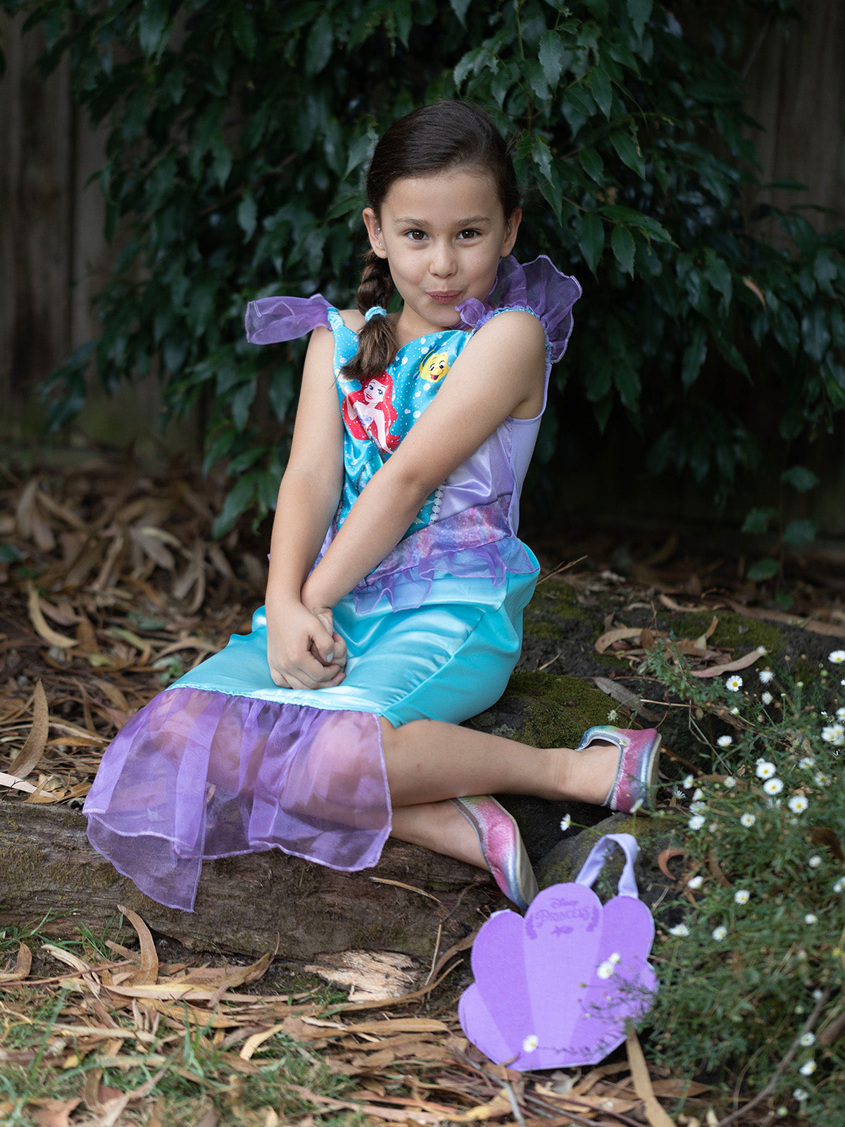 Girl sitting in the Ariel dress with purple tulle tail beside the purple shell-shaped handbag